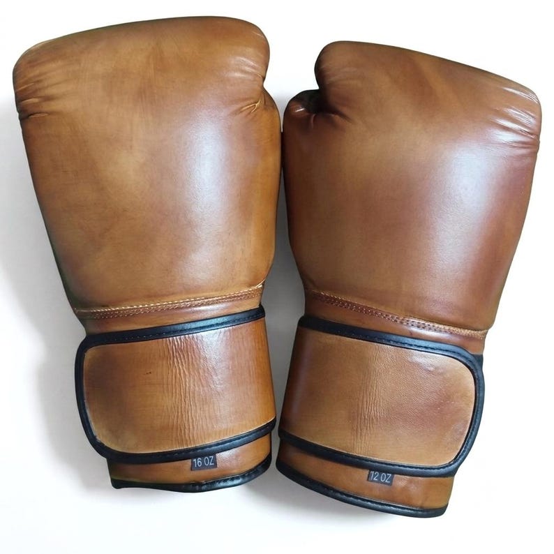 May include: A pair of tan leather boxing gloves with black trim. The left glove is labeled 16 oz and the right glove is labeled 12 oz. Both gloves feature a smooth, polished finish with visible stitching along the wrist and thumb areas.