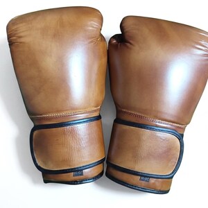 May include: A pair of tan boxing gloves featuring a smooth finish and black trim along the wrist straps. Each glove displays a small rectangular label at the base of the cuff with the text 10-OZ, indicating the weight in ounces for these items.