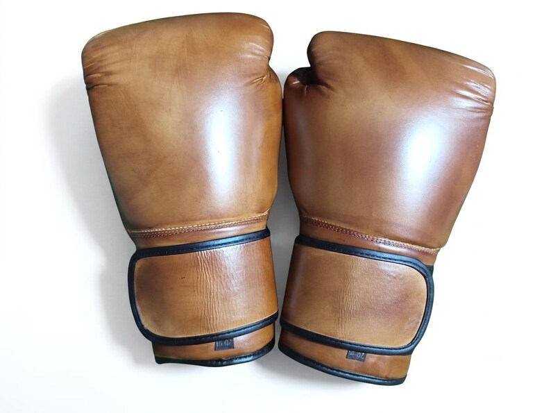 May include: A pair of tan boxing gloves featuring a smooth finish and black trim along the wrist straps. Each glove displays a small rectangular label at the base of the cuff with the text 10-OZ, indicating the weight in ounces for these items.