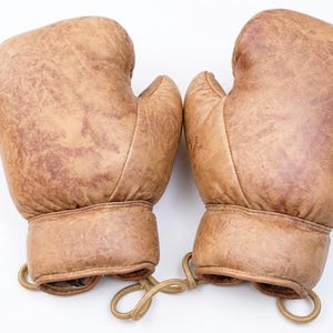 May include: A pair of vintage-style boxing gloves crafted from distressed tan leather. The gloves feature a smooth, rounded padding design with reinforced stitching at the wrist and a tan cord connecting the two gloves at the base of the cuffs.