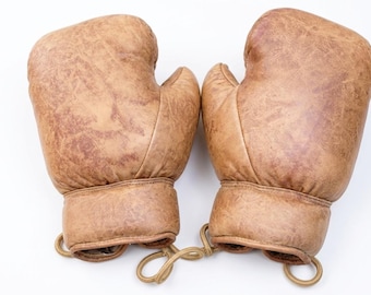 May include: A pair of vintage-style boxing gloves crafted from distressed tan leather. The gloves feature a smooth, rounded padding design with reinforced stitching at the wrist and a tan cord connecting the two gloves at the base of the cuffs.