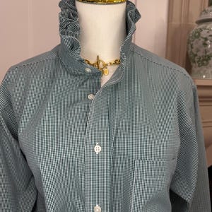May include: A long-sleeved button-down shirt in a dark green and white gingham check pattern. The shirt features a high, ruffled stand-up collar and a single chest pocket on the left side. The front is secured with white buttons.