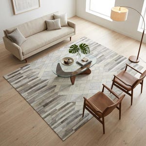 Op de afbeelding: Een rechthoekig vloerkleed met een geometrisch patchworkpatroon in grijze, witte en beige tinten. Het kleed ligt in een woonkamer met een beige bank, twee bruine leren stoelen en een ovale glazen salontafel.