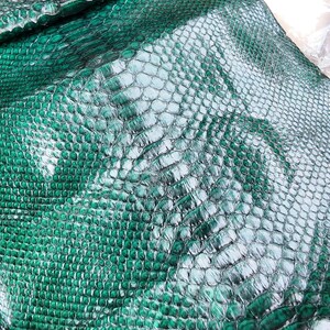 May include: A folded sheet of emerald green snakeskin leather featuring a glossy, textured scale pattern. The surface displays a natural, repeating diamond-shaped scale arrangement with a reflective finish. The text GOPYTHONHO is printed at the top.