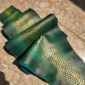 May include: A piece of python skin leather featuring a gradient of deep emerald green and metallic gold. The scales are textured and overlapping, displaying a natural pattern. The logo GoPythonHouse is printed at the top of the leather hide.