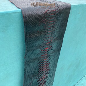 Puede incluir: Una pieza larga y rectangular de cuero de piel de serpiente negra con una capa inferior roja visible entre las escamas. El texto ExoTraditionalTannery est&aacute; impreso en una fuente decorativa en la parte superior central de la piel texturizada.