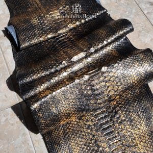 May include: A long, folded piece of genuine python skin leather featuring a metallic gold and black scale pattern. The surface displays a natural, textured reptilian scale design with a glossy finish. The text Gopythonhouse is printed at the top.