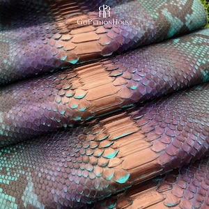 May include: A folded sheet of iridescent snakeskin leather featuring a gradient of metallic copper, deep purple, and vibrant teal scales. The surface displays a natural reptilian texture with the logo and text GOMPYTHONHOUSE printed at the top.
