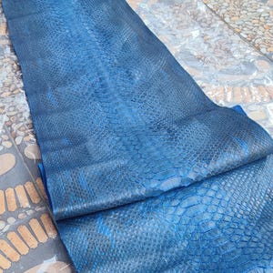 May include: A long, rectangular piece of blue python skin leather featuring a natural scale pattern with a gradient of dark and light blue tones. The surface displays a distinct texture with larger scales along the center and smaller scales on the sides. The text GoPythonHouse is printed in white near the top edge.