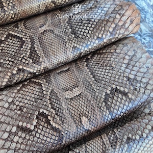 May include: A folded piece of natural snakeskin leather featuring a detailed scale pattern in shades of brown, tan, and cream. The surface displays a repeating diamond-like texture. The text GoPythonHouse is printed in white near the top center.