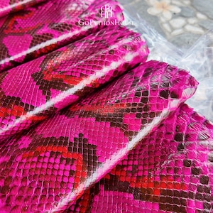 May include: A folded sheet of genuine python skin dyed in a vibrant hot pink color with natural dark brown and black scale patterns. The surface features a distinct reptilian texture with a glossy finish. The text GoPythonHouse is visible.