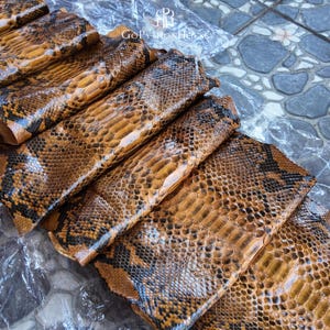 May include: A collection of natural python skin sheets featuring a distinct brown and black scale pattern. The textured reptile leather displays a glossy finish with overlapping scales. The text GoPythonHouse is visible on the plastic packaging.