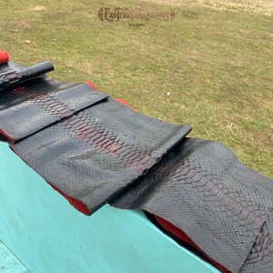 Puede incluir: Una tira larga y flexible de cuero de piel de serpiente negra con un patr&oacute;n de escamas distintivo y una parte inferior roja vibrante. El material est&aacute; doblado en secciones, revelando el interior rojo contrastante. El texto ExoTraditionalTannery es visible.