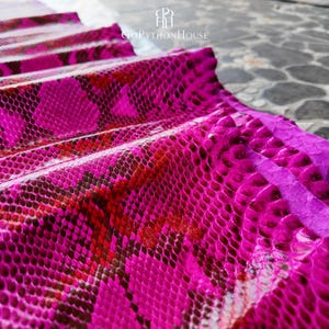 May include: A vibrant magenta and dark red snakeskin leather sheet with a glossy finish and a distinct scale pattern. The material is folded in layers, displaying a textured surface. The logo GoPythonHouse is visible at the top center.