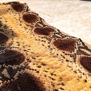 May include: A piece of natural snakeskin leather featuring a repeating pattern of dark brown and black diamond-shaped markings against a light tan and yellow background. The texture shows distinct, overlapping scales across the entire surface.
