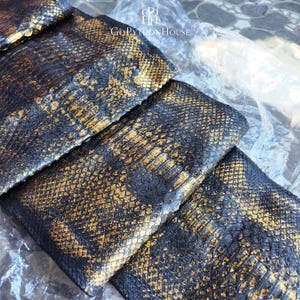 May include: A piece of textured python skin featuring a gradient of deep black and metallic gold scales. The surface displays a natural, repeating diamond pattern with a glossy finish. The logo GoPythonHouse is printed in white at the top center.