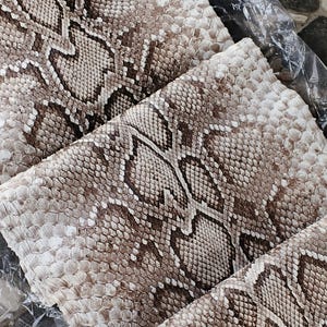 May include: A folded sheet of natural python skin featuring a repeating diamond pattern in shades of cream, beige, and dark brown. The surface displays a textured scale arrangement with the text GIO PYTHON HOUSE printed in white near the top edge.