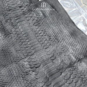 May include: A dark gray python skin with a distinct, overlapping scale pattern. The surface features a natural, textured finish with varying scale sizes. The text "GOPYTHONHOUSE" and a logo are printed in white at the top of the material.