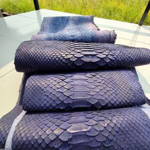 May include: A stack of three folded sheets of dark purple python skin leather with a distinct scale pattern. The top sheet is partially unfolded, revealing the texture. The logo GoPythonHouse is visible at the top center of the image.