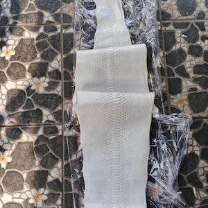 May include: A white python skin hide with a distinct scale pattern, folded into three sections and encased in a clear plastic sleeve. The top of the sleeve features the logo and text GUPYTHONHOUSE in white lettering against the textured skin surface.