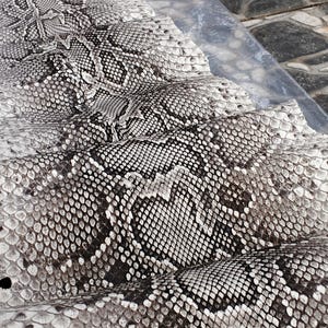 May include: A large, folded piece of natural python skin featuring a distinct black, white, and gray diamond-shaped scale pattern. The surface displays a textured, organic appearance with the text GoPythonHouse printed near the top edge.
