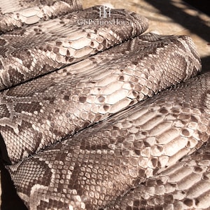 May include: A folded piece of natural python skin featuring a repeating scale pattern in shades of cream, beige, and dark brown. The surface displays a textured, organic appearance with a logo and the text GoPythonHouse printed at the top center.
