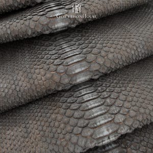 May include: A dark grey python skin leather hide with a natural scale pattern. The surface features a repeating texture of overlapping scales in varying sizes. The text GoPythonHouse is printed in white at the top center of the leather material.