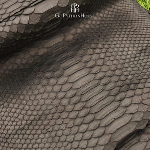 May include: A dark brown, textured python skin leather sheet featuring a natural scale pattern. The surface displays a gradient of scale sizes, from smaller scales on the edges to larger, elongated scales in the centre. The text GoPythonHouse is visible.