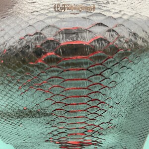 Puede incluir: Un material texturizado con un patr&oacute;n de escamas repetitivo en tonos verde azulado y negro, con una franja central prominente de acentos rojos vibrantes entre las escamas. El texto ExoTraditionalTannery est&aacute; impreso en la parte superior central.