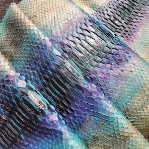 Könnte beinhalten: Ein Stück schillerndes Schlangenleder mit einem Farbverlauf aus metallisch blauen, violetten und türkisfarbenen Schuppen. Die Oberfläche zeigt ein natürliches, überlappendes Schuppenmuster mit reflektierendem Finish und dem Text PythonHouse.