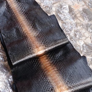 May include: A long, folded piece of black snakeskin leather featuring a prominent, light brown central stripe running down the length of the scales. The surface displays a detailed, repeating diamond-like texture. The text GoPythonHouse is visible.
