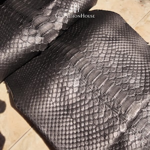 May include: A dark metallic gray python skin hide featuring a distinct natural scale pattern with a glossy finish. The text "GOPYTHONHOUSE" and a logo are printed in white at the top center of the textured, overlapping reptile leather surface.