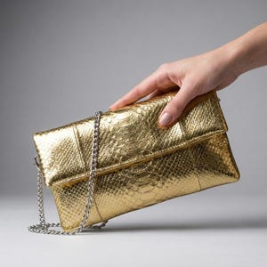 Puede incluir: Un bolso de mano dorado metálico con un patrón texturizado de piel de serpiente. El bolso rectangular tiene un diseño de solapa plegable e incluye una correa de hombro de cadena de metal plateado en los laterales.