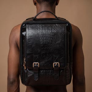 Può includere: Uno zaino nero caratterizzato da una texture a effetto pelle di coccodrillo. La borsa presenta un manico superiore, una patta frontale chiusa da due fibbie metalliche e uno scomparto laterale con cerniera verticale. Il design è di forma rettangolare.