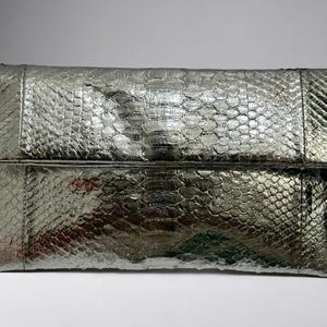 May include: A rectangular metallic silver clutch bag featuring a textured snakeskin pattern. The bag has a fold-over flap design with visible stitching along the edges and a glossy finish that reflects light across the surface of the material.