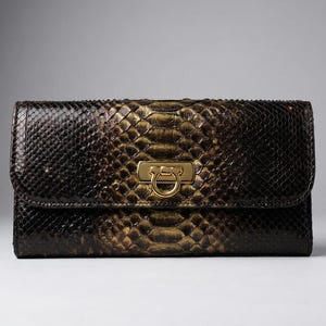 May include: A rectangular wallet featuring a dark brown and metallic gold snakeskin texture. The front flap is secured with a central gold-toned metal clasp that includes a circular ring detail. The wallet has a structured, horizontal orientation.