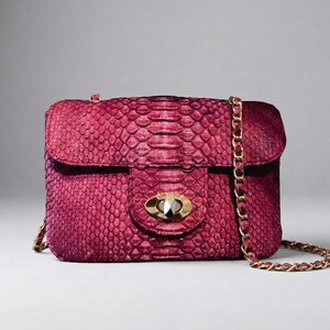 May include: A rectangular magenta handbag with a textured snakeskin pattern. The bag features a gold-tone metal turn-lock closure on the front flap and a matching gold-tone chain shoulder strap woven with magenta material for a cohesive look.