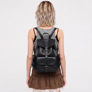 May include: A black leather backpack featuring a top flap with a buckle closure, a main compartment, and two side pockets. The bag includes adjustable shoulder straps and a top handle, showcasing a smooth finish and metal hardware accents.