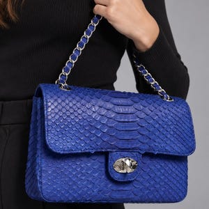 May include: A vibrant blue handbag featuring a textured snakeskin pattern, a rectangular flap closure, and a silver-toned metal turn-lock clasp. The bag includes a shoulder strap made of a silver metal chain interwoven with matching blue material.