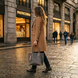 May include: A camel-coloured wool coat with a notched lapel and two buttons, paired with black trousers and brown leather ankle boots. A large, grey leather tote bag with a small pink tassel hangs from the side, featuring dual handles.