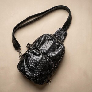 Pode incluir: Uma bolsa transversal preta com um padrão texturizado de pele de cobra. A bolsa possui um compartimento principal arredondado com fecho de zíper, um bolso frontal menor com zíper e uma alça de ombro ajustável com ferragens de metal prateado.