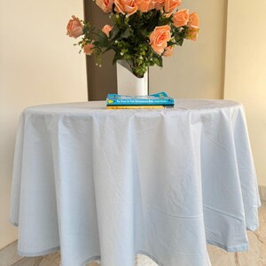May include: A round table covered with a solid light blue tablecloth. On the table sits a white vase filled with a bouquet of peach-colored roses and green foliage, along with two stacked books with blue and yellow covers.