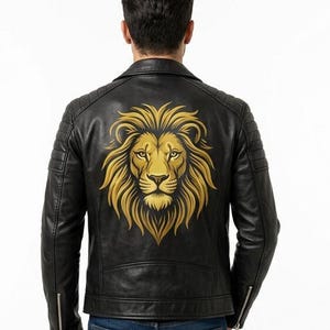 Könnte beinhalten: Eine schwarze Lederjacke mit einem großen, detaillierten goldenen Löwenkopf-Motiv auf dem Rücken. Die Jacke verfügt über einen klassischen Kragen, gesteppte Schulterpartien und silberfarbene Metallreißverschlüsse an den Ärmeln, getragen über einer blauen Jeans.