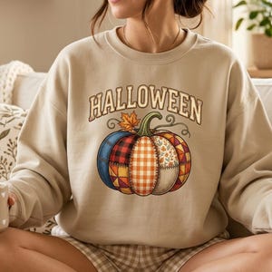 以下が含まれることがあります： ベージュ色のクルーネックのスウェットシャツ。アーチ状の茶色の文字で「HALLOWEEN」と書かれ、その下に大きなカボチャのグラフィックが描かれています。カボチャはチェック柄、ギンガムチェック、花柄など、さまざまな色の布のパッチワークで構成されており、緑色の茎が付いています。