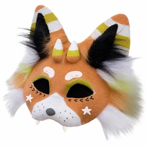 May include: A handmade fox mask with orange, white, black, and yellow accents. The mask features candy corn-shaped horns, star details, and black mesh eye coverings. The ears have striped patterns, and the mask has fluffy white and yellow fur.
