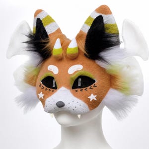 May include: A handcrafted fox mask with orange, white, black, and yellow accents. The mask features white star details, black mesh eyes, and small white fangs. The ears have striped patterns and the mask has white fluffy accents.