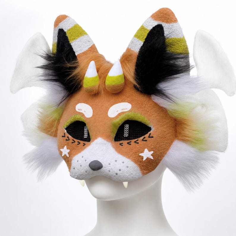 May include: A handcrafted fox mask with orange, white, black, and yellow accents. The mask features white star details, black mesh eyes, and small white fangs. The ears have striped patterns and the mask has white fluffy accents.