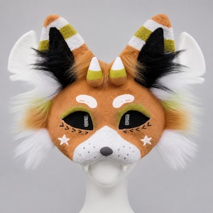 May include: A handcrafted fox mask with orange, white, black, and green accents. The mask features a white muzzle, black mesh eye coverings, and decorative star and arrow details. The ears have candy corn-like tips and striped patterns.
