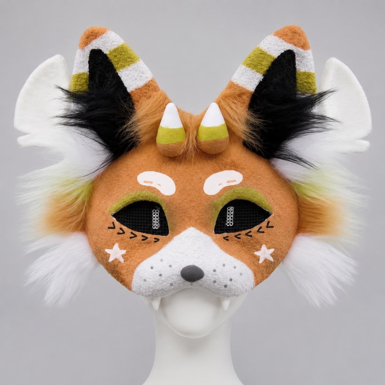 May include: A handcrafted fox mask with orange, white, black, and green accents. The mask features a white muzzle, black mesh eye coverings, and decorative star and arrow details. The ears have candy corn-like tips and striped patterns.
