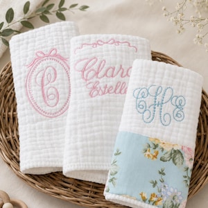 May include: Three white baby washcloths with embroidered details. One cloth features a pink letter "C" within a circle, another has the name "Clara Estelle" in pink, and the third has blue initials. The third cloth has a floral pattern.